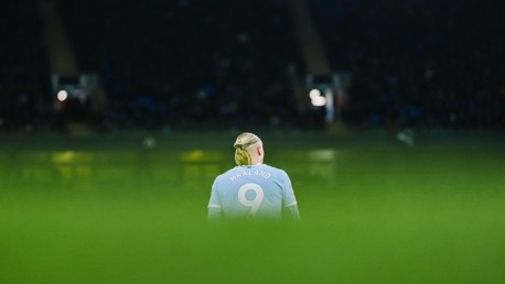 A football player with the name 'Haaland' and the number 9 on a light blue jersey stands on a field with blurred green foreground and dark background.