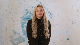 A person with long wavy hair, wearing a black sweater, standing against a textured blue and beige wall.