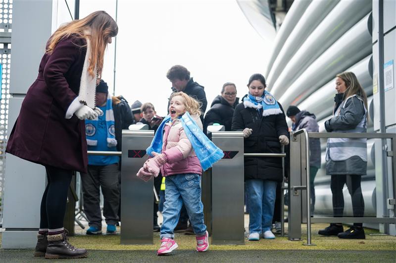 A child wearing pink and blue clothes stands at the entrance of a stadium, looking at an adult woman, with more people queued behind at the turnstiles.