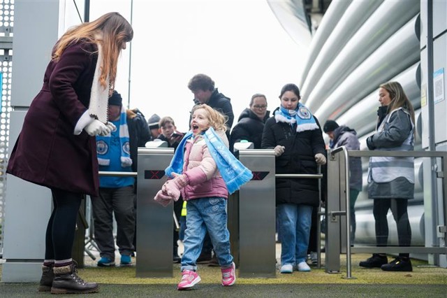 A child wearing pink and blue clothes stands at the entrance of a stadium, looking at an adult woman, with more people queued behind at the turnstiles.