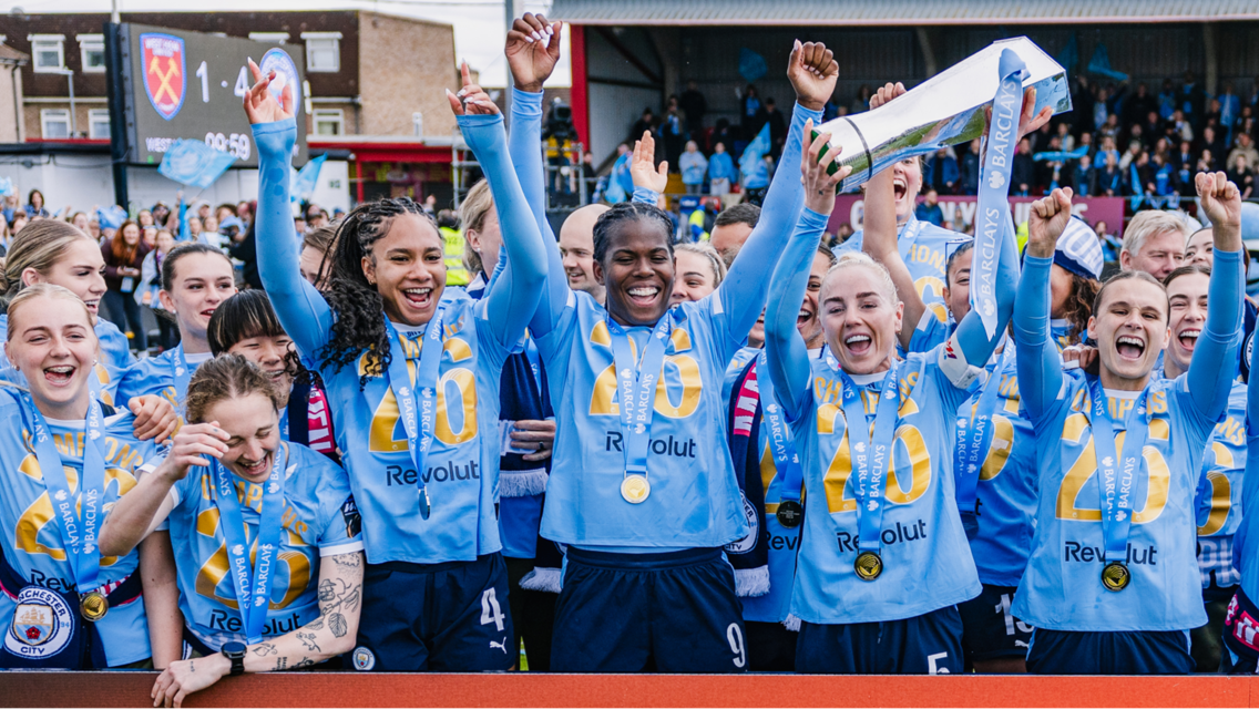 Manchester City Women's team celebrating with a trophy, wearing medals and team shirts. The scoreboard shows a score of West Ham 1-4 Manchester City.