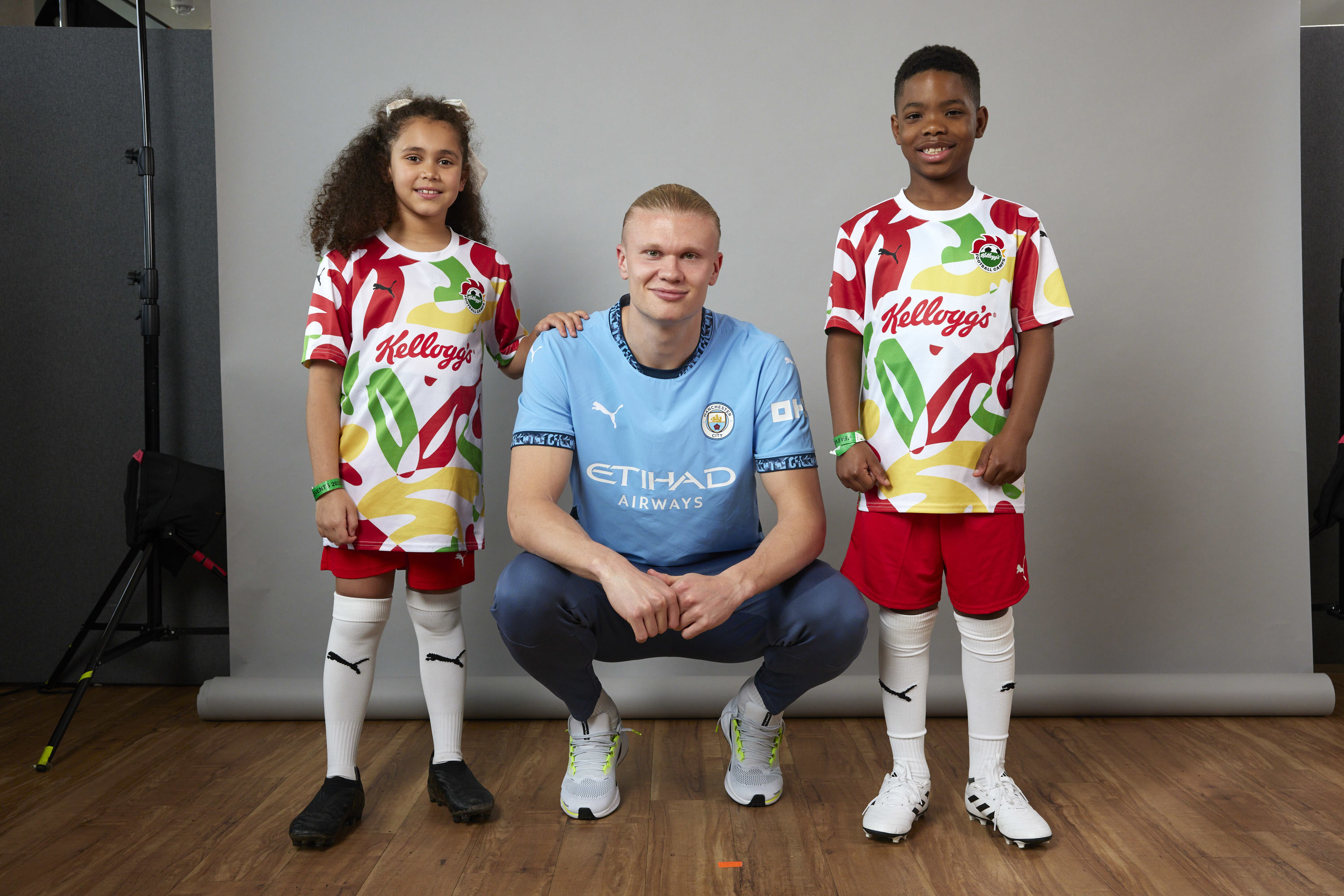 An image of a Manchester City player wearing the team's blue jersey, squatting between two mascots in Kellogg's-branded uniforms, with faces blurred.