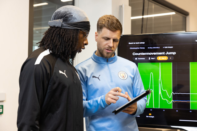 Two individuals in Puma sportswear discussing data on an electronic device. A screen displays 'Countermovement Jump.' One person wears a Manchester City jacket.