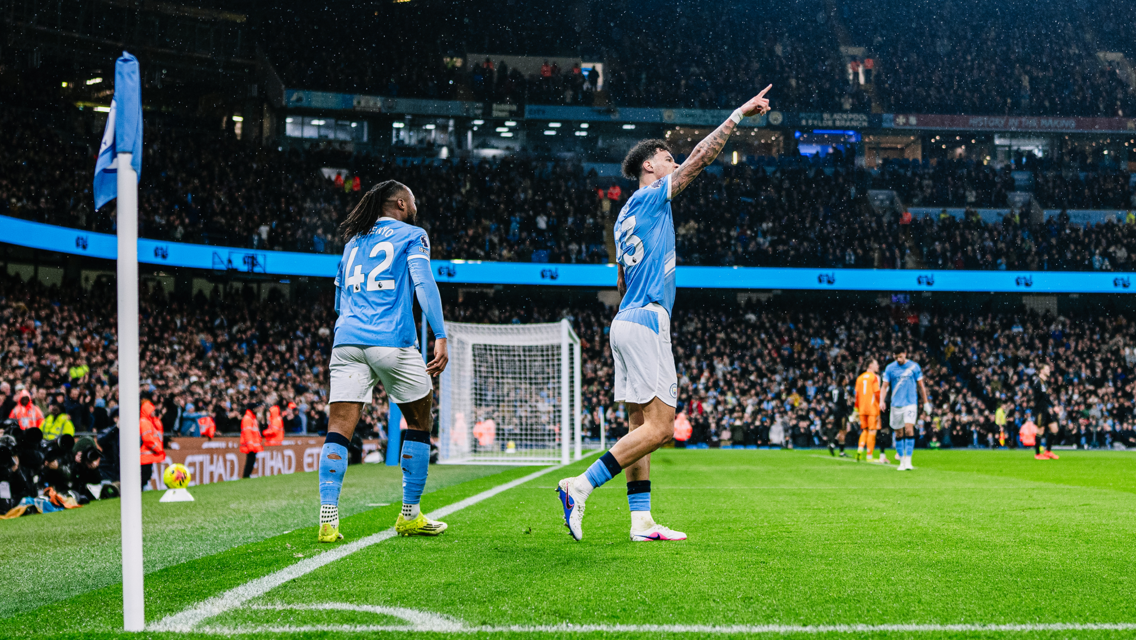 Football players celebrating a goal at a stadium, with a player pointing and another player standing near a corner flag wearing a Manchester City kit.