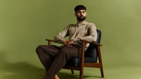 A man with a beard is sitting on a wooden chair with a green background. He is wearing a beige sweater and brown pants.