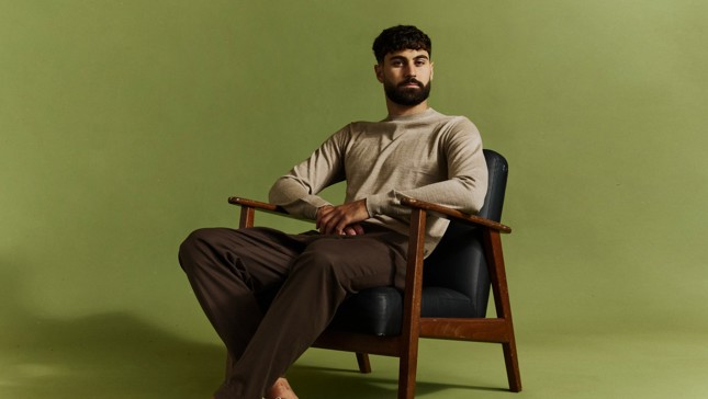 A man with a beard is sitting on a wooden chair with a green background. He is wearing a beige sweater and brown pants.