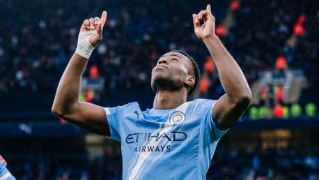 A Manchester City player with a blurred face is celebrating on the field, raising both arms with index fingers pointing upwards. The player's jersey features the Manchester City logo and Etihad Airways branding.