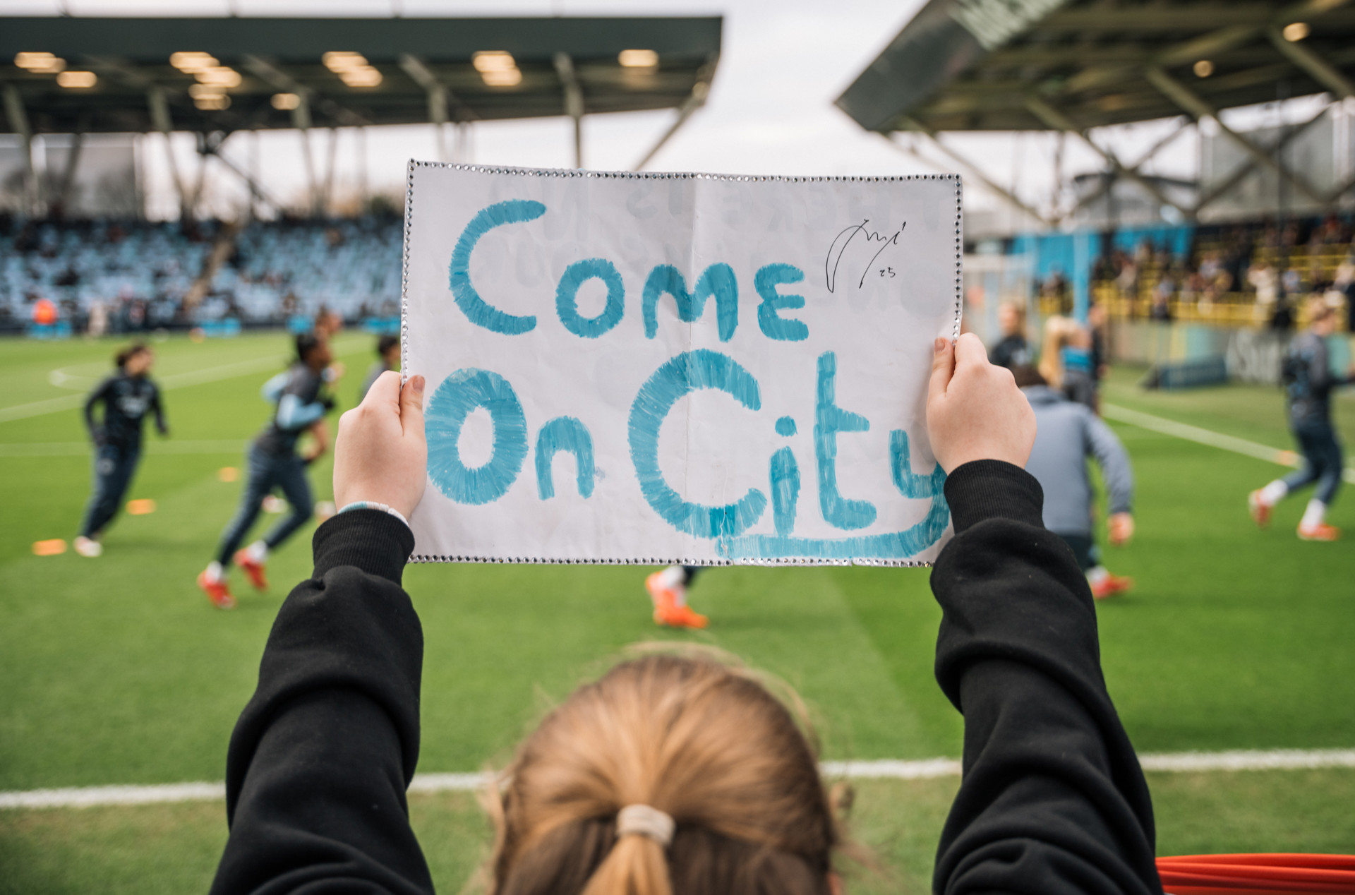 A person holds a sign saying 'Come On City' with a Manchester City signature at a football match, with players warming up in the background.