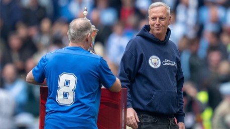 Two men at a Manchester City community event, one wearing a Man City hoodie and the other holding a trophy.