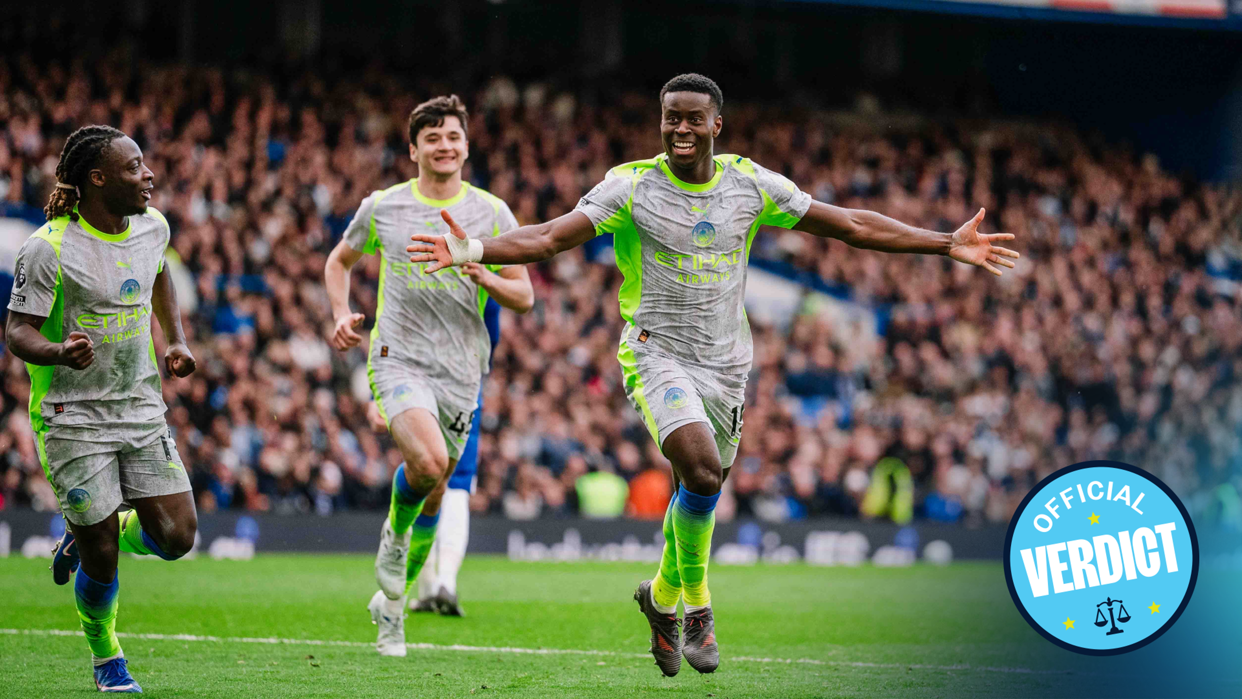 Manchester City players celebrating a goal on the field with the crowd in the background.