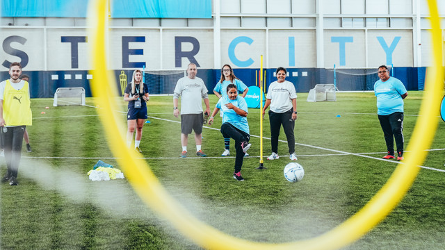 A group of people, possibly trainers and participants, are engaged in a football training session at an indoor facility with 'MANCHESTER CITY' written on the wall.