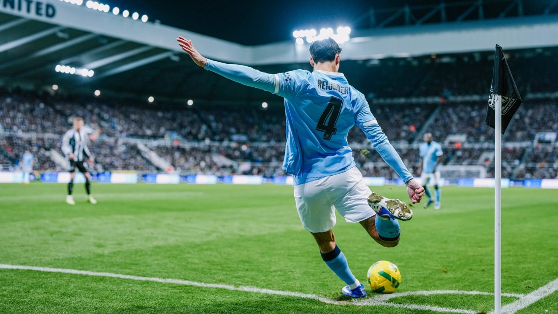 A soccer player from Manchester City in a light blue jersey (number 4) takes a corner kick during an evening match at a stadium with bright floodlights.