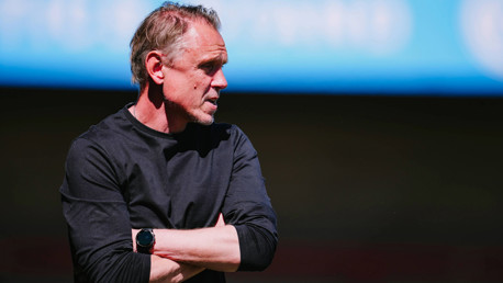 A man in a black long-sleeve shirt stands with arms crossed, wearing a black smartwatch. The background is a blurred blue sky and dark ground.