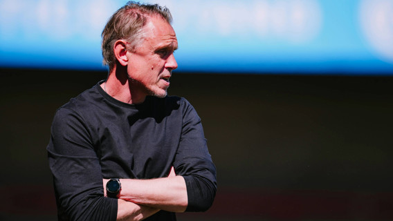 A man in a black long-sleeve shirt stands with arms crossed, wearing a black smartwatch. The background is a blurred blue sky and dark ground.