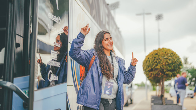 A person wearing a blue jacket and lanyard stands outside a bus with blurred face, raising both hands pointing upward. The bus has logos for Etisalat and Midea visible on it.