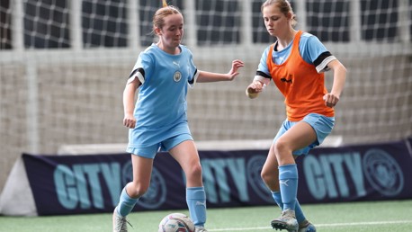 Two young soccer players, one in a blue kit and another wearing an orange bib, are playing on a football pitch with a 'CITY' banner in the background.