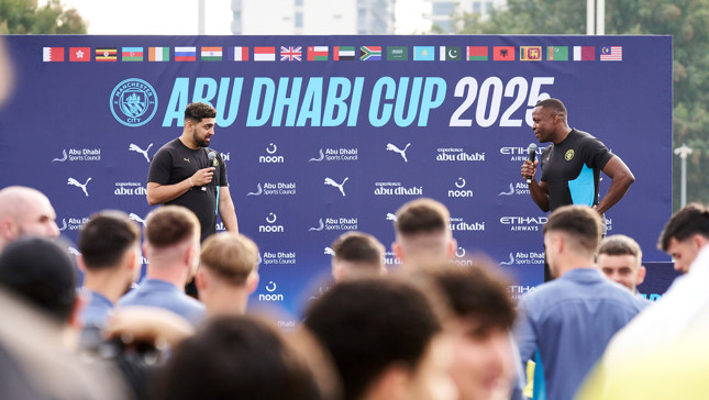 Two individuals are speaking at an event in front of a banner that reads 'Abu Dhabi Cup 2025' with multiple international flags displayed above. The banner includes Manchester City logos and sponsors such as Puma, Etihad Airways, and Noon.