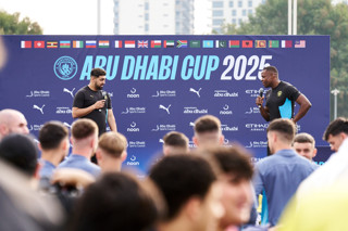 Two individuals are speaking at an event in front of a banner that reads 'Abu Dhabi Cup 2025' with multiple international flags displayed above. The banner includes Manchester City logos and sponsors such as Puma, Etihad Airways, and Noon.