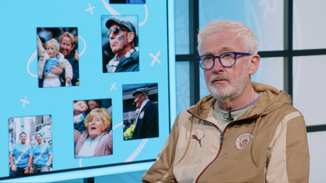 A person in a brown Manchester City Puma jacket is seated beside a display featuring a collage of various blurred photos, possibly of fans or events.