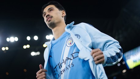 A Manchester City player holding open a light blue jacket to reveal the team jersey with the club crest and sponsor logo visible.