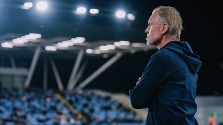 A coach wearing a navy hoodie stands with arms crossed under stadium lights, watching a match. Blurred seating is visible in the background.