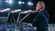 A coach wearing a navy hoodie stands with arms crossed under stadium lights, watching a match. Blurred seating is visible in the background.