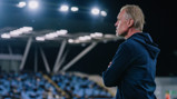 A coach wearing a navy hoodie stands with arms crossed under stadium lights, watching a match. Blurred seating is visible in the background.