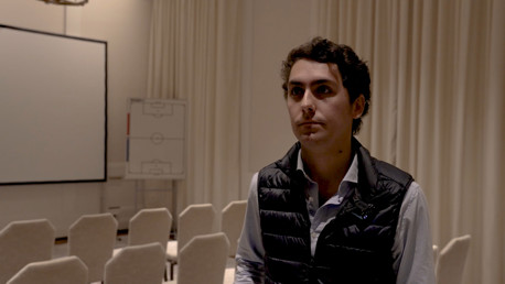 A person in a black vest stands in a meeting room with chairs, a projection screen, and a soccer strategy board in the background.