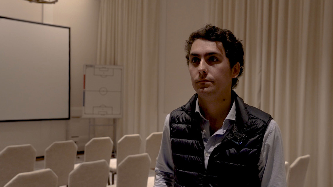 A person in a black vest stands in a meeting room with chairs, a projection screen, and a soccer strategy board in the background.