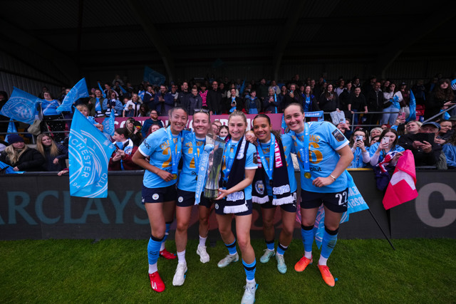 Group of Manchester City Women players celebrating with a trophy on the field, with fans cheering in the background. Players are wearing blue jerseys and medals.