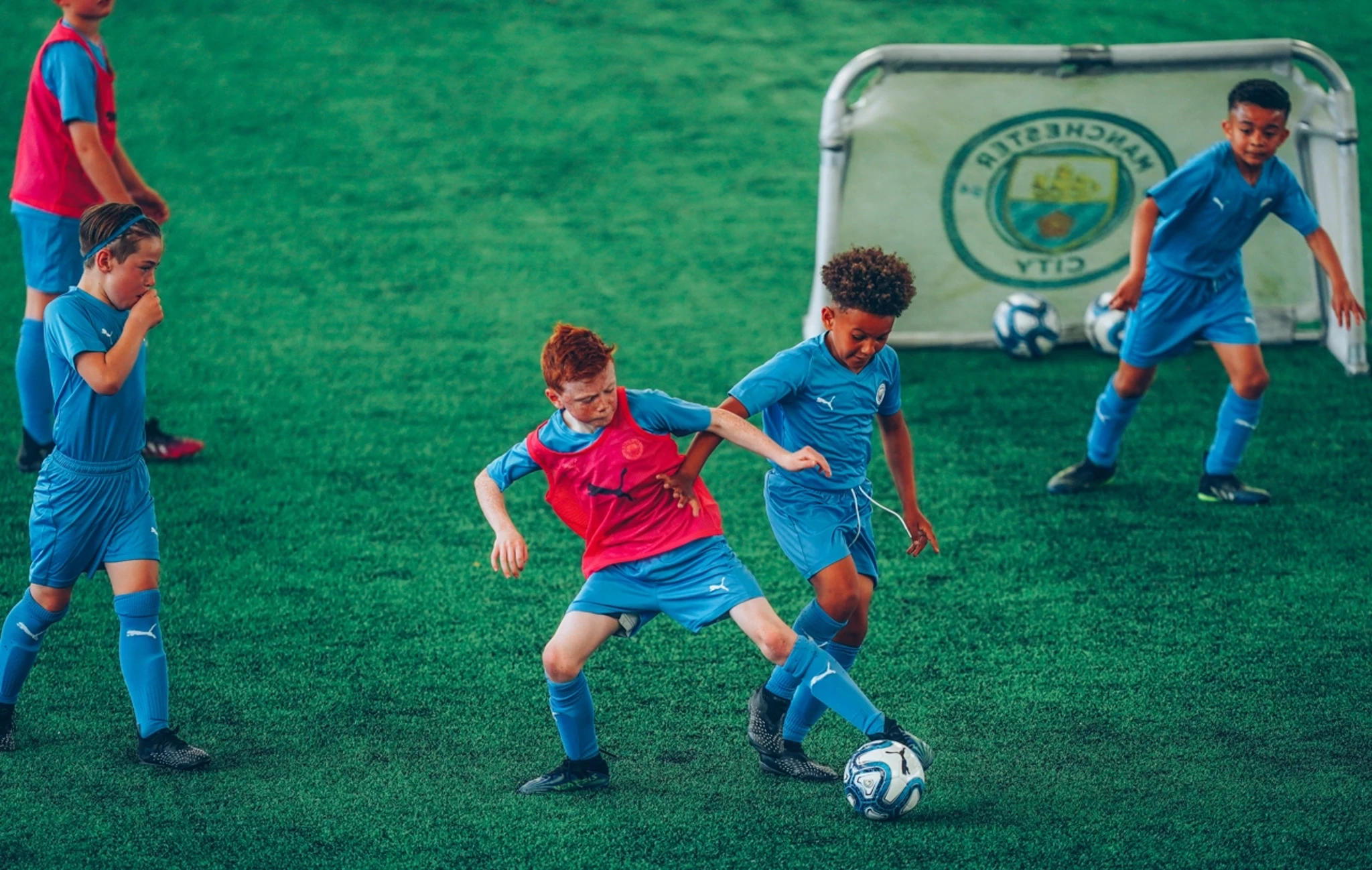 Children playing soccer on an artificial turf field. One child is wearing a red training bib and the others are in blue uniforms. A small goalpost with the Manchester City logo is in the background.