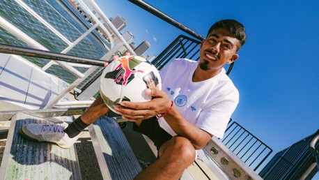 A person sitting on outdoor stairs holding a Puma soccer ball, wearing a white shirt with blue logo, with a waterfront view and city skyline in the background.