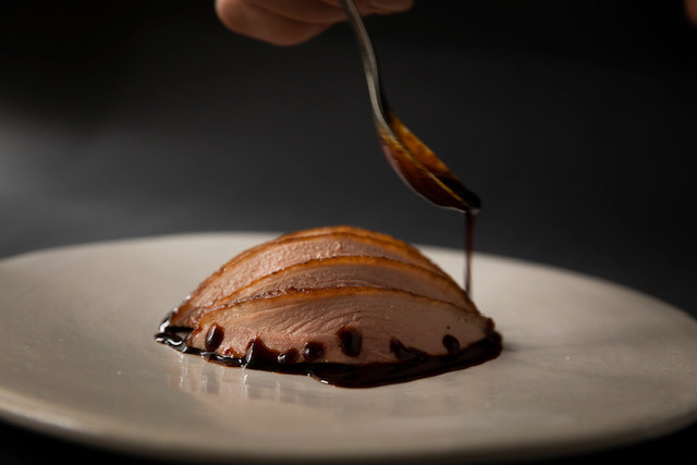 A hand delicately drizzles sauce over thinly sliced meat presented on a white plate.