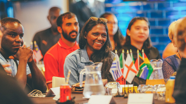 A diverse group of people sit around a table with various national flags on it, including Malaysia, Greece, and South Africa. A soda can and water jug are also visible on the table.