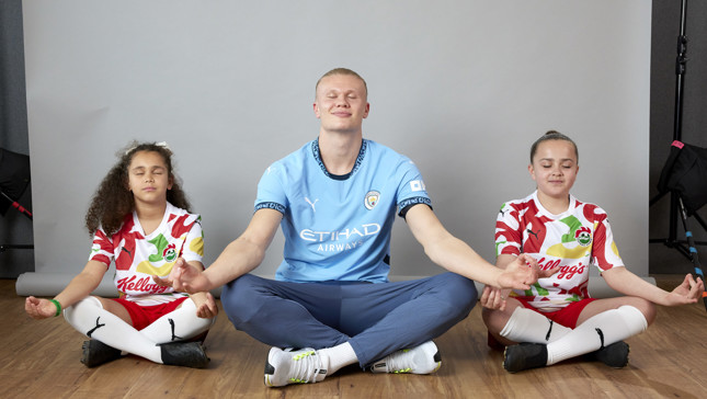 A Manchester City player is sitting in a meditation pose, flanked by two children wearing soccer jerseys. The scene is indoors, with a neutral background.