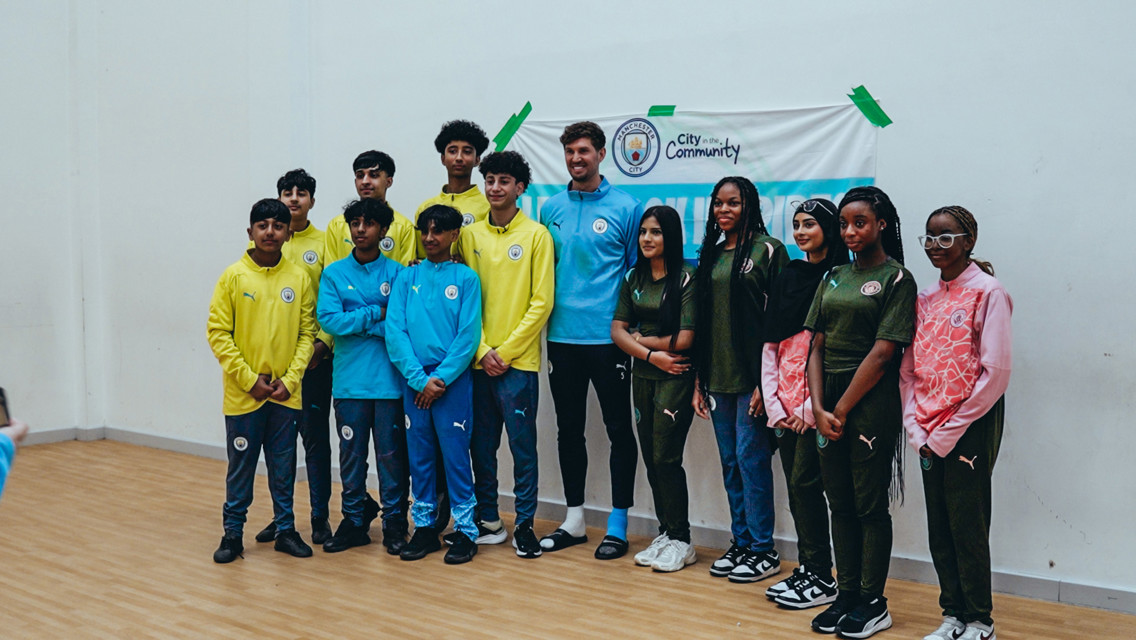 Group photo of young people and an adult wearing Manchester City attire at a community event, with a banner in the background.