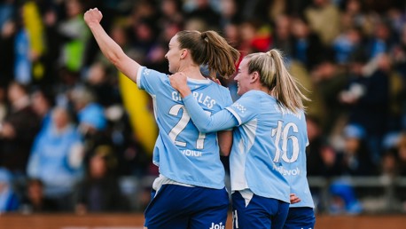 Three soccer players in light blue jerseys celebrate a goal. One player has their arm raised. The jerseys display the numbers 21 and 16, with names and sponsorship logos visible.