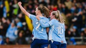 Three soccer players in light blue jerseys celebrate a goal. One player has their arm raised. The jerseys display the numbers 21 and 16, with names and sponsorship logos visible.