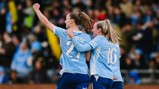 Three soccer players in light blue jerseys celebrate a goal. One player has their arm raised. The jerseys display the numbers 21 and 16, with names and sponsorship logos visible.