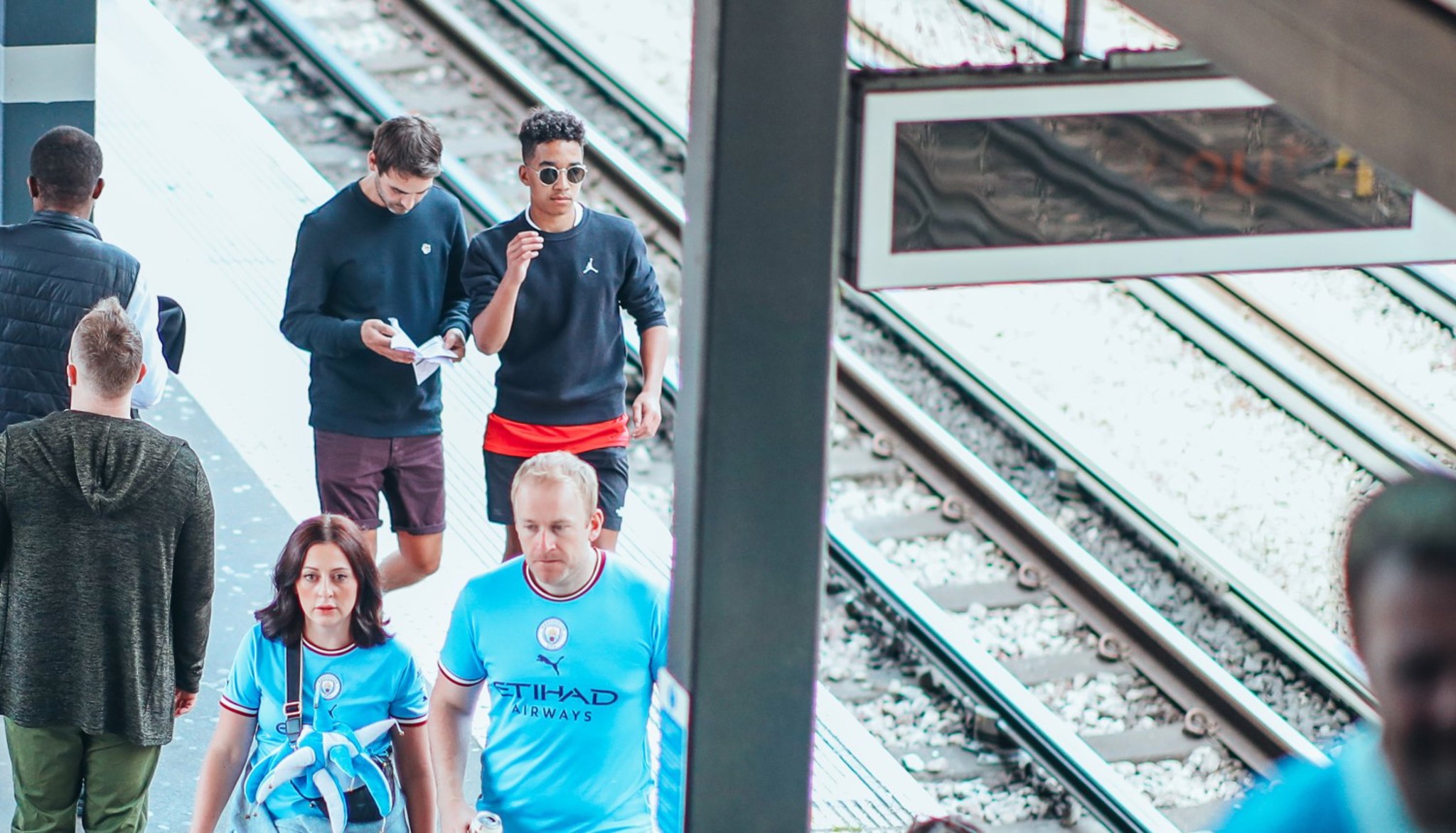 People at a train station, some wearing Manchester City jerseys, with faces blurred for privacy.