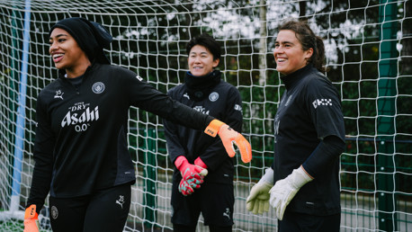 Three individuals wearing black training gear with Manchester City and Asahi logos, standing in front of a goal net. They are wearing goalkeeper gloves.