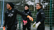 Three individuals wearing black training gear with Manchester City and Asahi logos, standing in front of a goal net. They are wearing goalkeeper gloves.