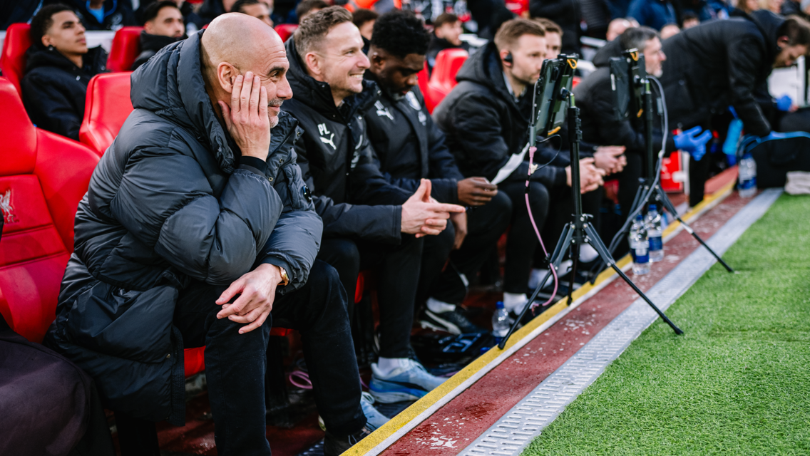 Soccer team staff seated on a bench trackside, wearing dark jackets, communicating with hand gestures.