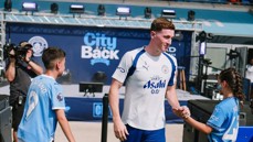 A Manchester City player interacts with young fans at an event. Background features promotional material for City and sponsor Asahi.