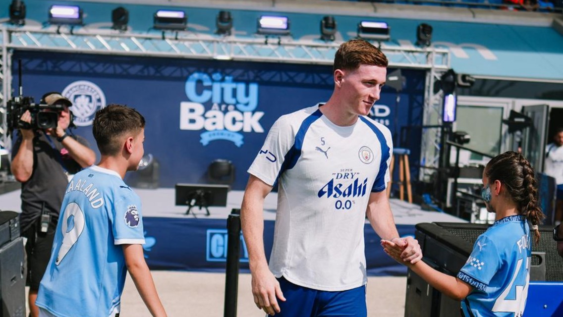 A Manchester City player interacts with young fans at an event. Background features promotional material for City and sponsor Asahi.