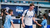 A Manchester City player interacts with young fans at an event. Background features promotional material for City and sponsor Asahi.