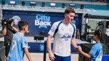 A Manchester City player interacts with young fans at an event. Background features promotional material for City and sponsor Asahi.