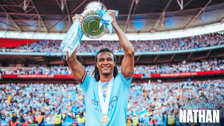 A football player in a blue jersey holds up a silver trophy with blue ribbons labeled 'Emirates FA Cup'. Text says 'Thank You Nathan'. The background shows a large crowd in a stadium.