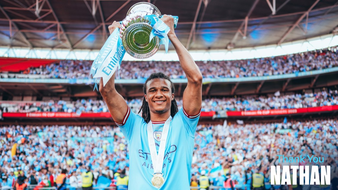 A football player in a blue jersey holds up a silver trophy with blue ribbons labeled 'Emirates FA Cup'. Text says 'Thank You Nathan'. The background shows a large crowd in a stadium.