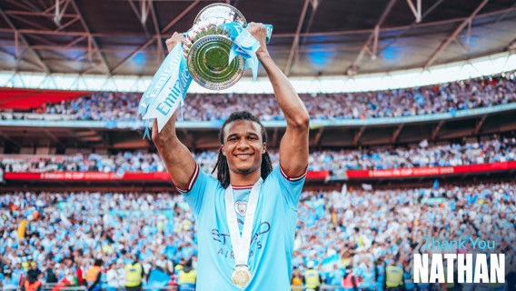 A football player in a blue jersey holds up a silver trophy with blue ribbons labeled 'Emirates FA Cup'. Text says 'Thank You Nathan'. The background shows a large crowd in a stadium.
